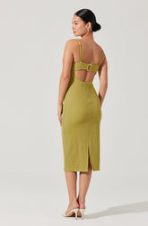 A woman wearing a fitted, sleeveless dress in a green geometric pattern. The dress features adjustable shoulder straps, a wide waistband with a metallic buckle, and a cutout detail at the back with a small slit at the hem. She is also wearing light-colored high-heeled shoes.